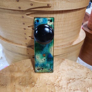 以下が含まれることがあります： 緑と青の塗装が施された金属製のバックプレートが付いた黒いドアノブ。バックプレートは使い古したような外観です。