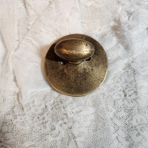 May include: A vintage brass cabinet knob with an oval handle and a circular base. The knob has a tarnished, aged appearance, suggesting it is antique. The background is a white lace fabric.
