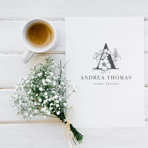 May include: A white card with a black floral design of the letter A and the text "Andrea Thomas Floral Designs". A cup of coffee and a bouquet of white flowers are on a white wooden surface.