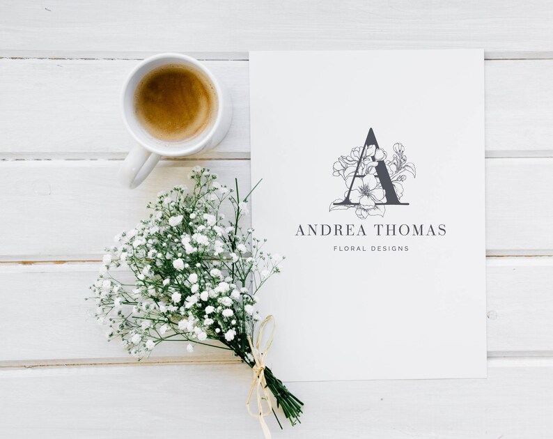 May include: A white card with a black floral design of the letter A and the text "Andrea Thomas Floral Designs". A cup of coffee and a bouquet of white flowers are on a white wooden surface.