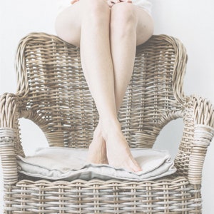 May include: A person sits in a wicker chair with a white cushion. The chair is made of natural woven wicker and has a light brown color. The person's legs are crossed and their feet are resting on the cushion. The background is a white wall with the text "WILD THREADS - organic clothing" printed on it.