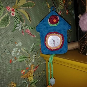 May include: A crocheted cuckoo clock with a blue house, red roof, and a yellow, orange, and red clock face. The clock has a green, yellow, and orange pendulum with a light blue and orange ball at the end.