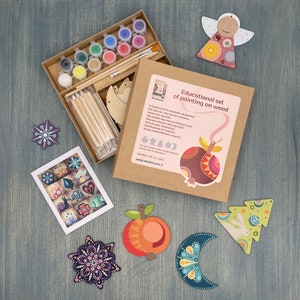 May include: A craft kit for painting wooden ornaments. The kit includes paints, brushes, wooden shapes, and instructions. The shapes include a crescent moon, a star, a tree, a peach, and an angel. The box is brown with a colourful pomegranate design and the text "Educational set of painting on wood".