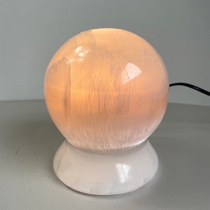 May include: A white ceramic table lamp with a round, frosted glass shade. The lamp is lit and casts a warm glow.