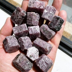 May include: A collection of rough, purple ruby crystals in various sizes and shapes. The crystals have a natural, unpolished look and are arranged in a pile on a hand.