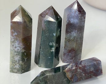 May include: Four polished gemstone obelisks. The stones feature a mix of colors, including deep red, green, and white. The obelisks have a rectangular base and a pointed top. The stones are arranged on a white surface.
