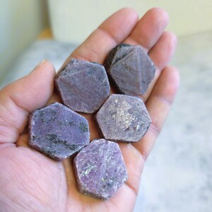 May include: Five rough-cut, hexagonal ruby gemstones in varying shades of purple and pink are held in a hand. The stones have a natural, unpolished appearance, with visible textures and subtle colour variations. The gemstones are displayed against a neutral background.