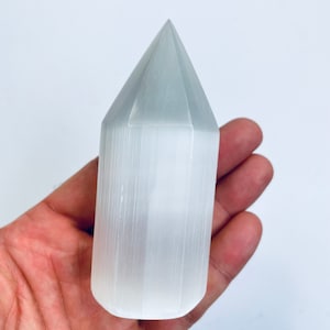 May include: A white selenite crystal point, also known as a selenite wand, is held in a hand. The crystal is smooth and has a pointed tip. Selenite is a type of gypsum.