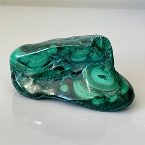 May include: A polished malachite stone with a swirling pattern of dark and light green hues. The natural stone has a smooth, glossy surface and a unique, organic shape. This decorative stone is a beautiful addition to any collection.