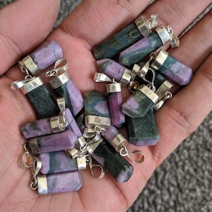 May include: A collection of 15 purple and green gemstone pendants with silver bails. The pendants are all rectangular in shape and have a polished finish.