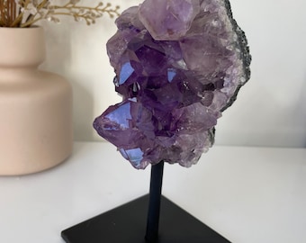 May include: A large amethyst crystal cluster on a black metal stand. The crystal is a deep purple color and has a natural, rough texture.
