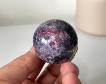May include: A polished purple and pink sphere of lepidolite, a type of lithium mica, with a smooth, shiny surface. The sphere is about the size of a golf ball and is held in a hand.