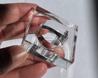May include: A clear acrylic ring holder with a square base. The holder is designed to hold a ring securely in place.