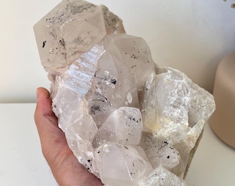 May include: A large, clear quartz crystal cluster with black inclusions. The crystal is translucent and has a rough, natural texture.