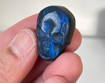 Puede incluir: Un cráneo tallado en un cristal de labradorita azul. El cristal tiene un brillo iridiscente.