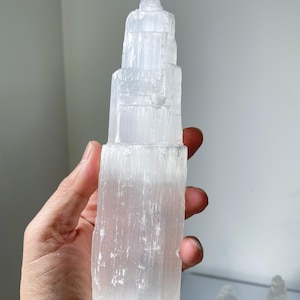 May include: A clear selenite tower with a layered design. The tower is held in a hand and is made up of stacked, cylindrical sections. The surface has a textured, crystalline appearance. The background is a neutral, off-white colour.