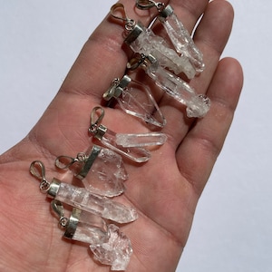 Pode incluir: Uma coleção de sete pingentes de cristal de quartzo transparente com engastes de prata. Os cristais são de várias formas e tamanhos, alguns são longos e finos, outros são mais curtos e largos.