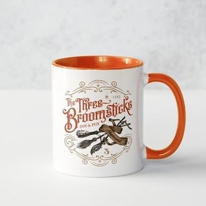 May include: A white ceramic mug with an orange rim and handle. The mug features a graphic of the "Three Broomsticks" pub from the Harry Potter series, with the text "The Three Broomsticks" and a drawing of three broomsticks.
