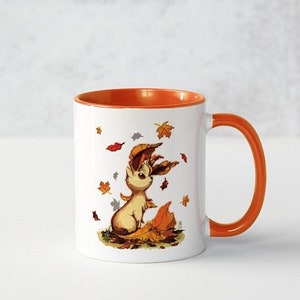 Op de afbeelding: Een witte keramische mok met een oranje rand en handvat. De mok heeft een cartoonillustratie van een bruine en witte vos met herfstbladeren eromheen.