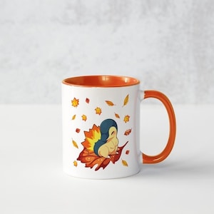 May include: A white ceramic mug with an orange rim and handle. The mug features a cartoon illustration of a small, blue and orange creature sitting on a pile of autumn leaves. The background is white with scattered orange and yellow leaves.