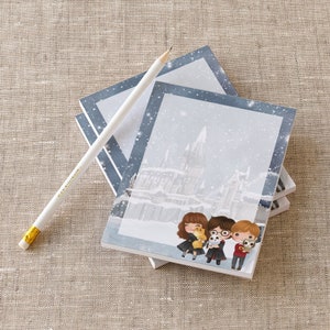 May include: A stack of blue and white notepad paper with a snowy scene of Hogwarts castle and Harry Potter characters. The characters are holding their animal companions. The notepad paper is on a beige fabric surface.