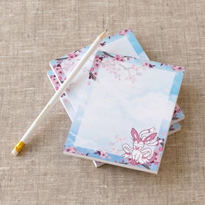 May include: A stack of three pink and blue notepad sets with a pink and white cartoon character on the front. The notepad has a blue background with white clouds and pink cherry blossoms.