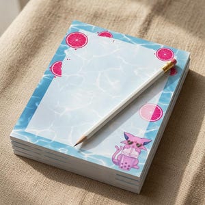 May include: A rectangular notepad featuring a light blue water-themed border with pink grapefruit slices and a small purple cat character wearing heart-shaped sunglasses while holding a boba tea drink, accompanied by a white wooden pencil.