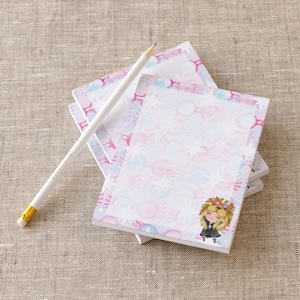 May include: A stack of pink and blue patterned notepad sets with a cartoon illustration of a girl wearing a wreath and glasses. Each notepad has a blank white space for writing.