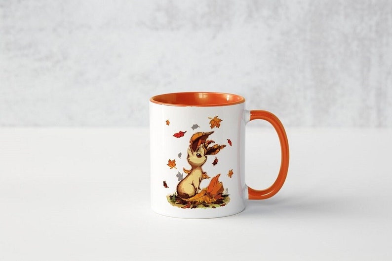 May include: A white ceramic mug with an orange rim and handle. The mug features a cartoon illustration of a brown and white fox with autumn leaves around it.