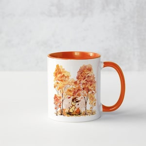 May include: A white ceramic mug with an orange rim and handle. The mug features a watercolour illustration of a small animal sitting in a forest of orange and yellow trees.