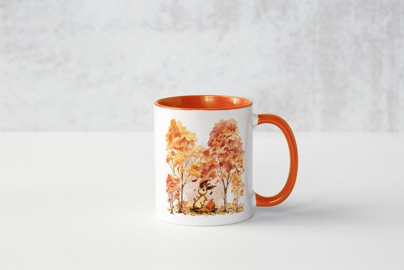 May include: A white ceramic mug with an orange rim and handle. The mug features a watercolour illustration of a small animal sitting in a forest of orange and yellow trees.