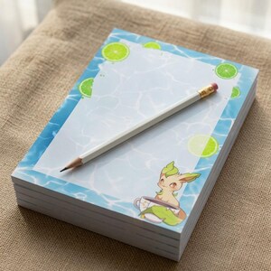 May include: A rectangular notepad with a light blue water-themed border featuring lime slice illustrations. The bottom right corner displays a cartoon creature sitting in a teacup. A white wooden pencil with a pink eraser rests on the notepad.