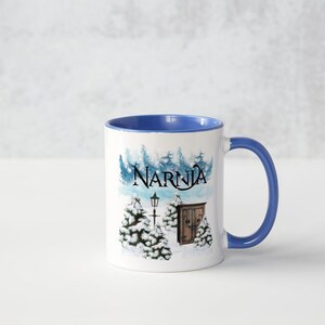 May include: A white ceramic mug with a blue rim and handle. The mug features a winter scene with a lamppost, a wooden door, and trees covered in snow. The text "Narnia" is printed in black on the mug.