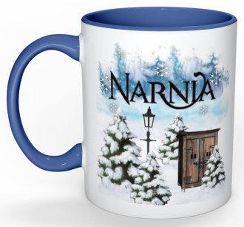 May include: White ceramic mug with a blue rim and handle. The mug features a winter scene with snow-covered trees, a lamppost, and a wooden door with the word "Narnia" written in a stylized font.