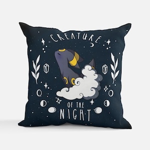 May include: Dark blue decorative pillow with a cartoon creature design. The pillow features a stylized creature with yellow accents, a white cloud, and the text "CREATURE OF THE NIGHT" in white. The design includes stars, moons, and crystals.