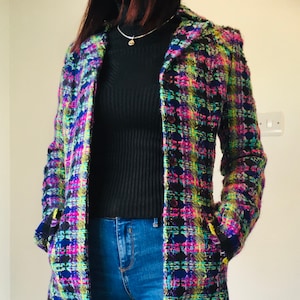 May include: A long-sleeved blazer featuring a vibrant multicolored plaid pattern in neon pink, yellow, blue, and black. The jacket has a textured tweed-like finish, a notched collar, front pockets, and a button-down front closure, worn over a black top.