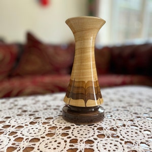 May include: A wooden vase with a brown and tan striped design. The vase is sitting on a white crocheted tablecloth.