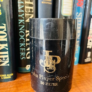 May include: A black cylindrical container with gold lettering that reads "John Player Special 50 FILTER." The container is in front of a collection of books with various titles and colors.