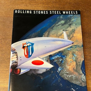 May include: A black and white concert program for the Rolling Stones Steel Wheels tour in Tokyo, 1990. The cover features a white airplane flying over a blue and green globe with a red circle on the tail.