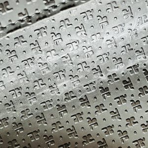 May include: Close-up of a textured, dark gray surface with a repeating pattern of stylized letter "D" shapes and small dots. The pattern is embossed, creating a subtle relief effect. The surface appears to be made of a durable material.