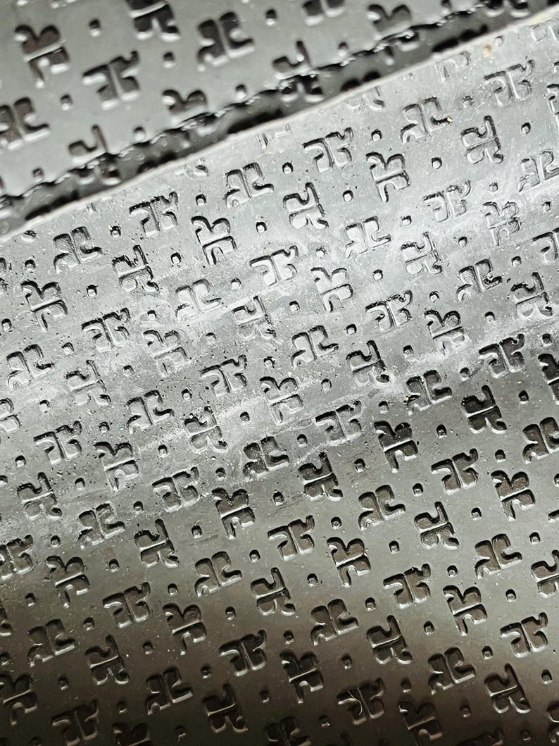 May include: Close-up of a textured, dark gray surface with a repeating pattern of stylized letter "D" shapes and small dots. The pattern is embossed, creating a subtle relief effect. The surface appears to be made of a durable material.