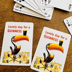 May include: Playing cards with a toucan illustration and the text "Lovely day for a GUINNESS." A fan of playing cards is visible, with the suits of clubs and spades. The cards are on a wooden surface.