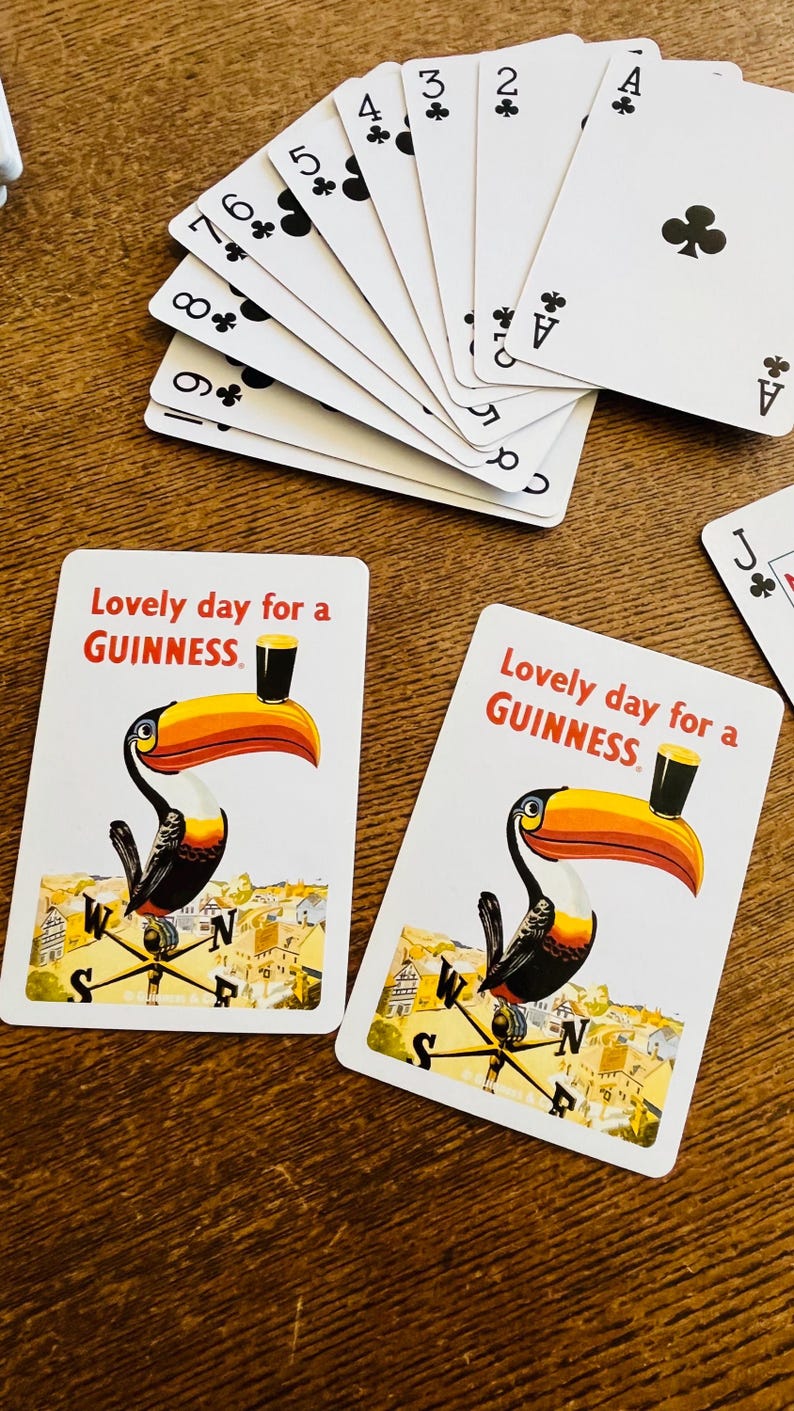 May include: Playing cards with a toucan illustration and the text "Lovely day for a GUINNESS." A fan of playing cards is visible, with the suits of clubs and spades. The cards are on a wooden surface.
