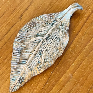 May include: A silver-toned, leaf-shaped ornament with a textured finish, featuring gold and blue accents. The leaf's detailed veining is visible. It is displayed on a wooden surface. The object is likely a decorative item.