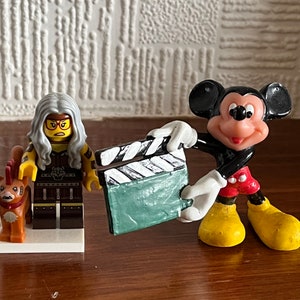 May include: A black and white clapperboard held by a red and yellow Mickey Mouse figurine. A Lego figurine with grey hair and a yellow and brown outfit stands next to Mickey Mouse.