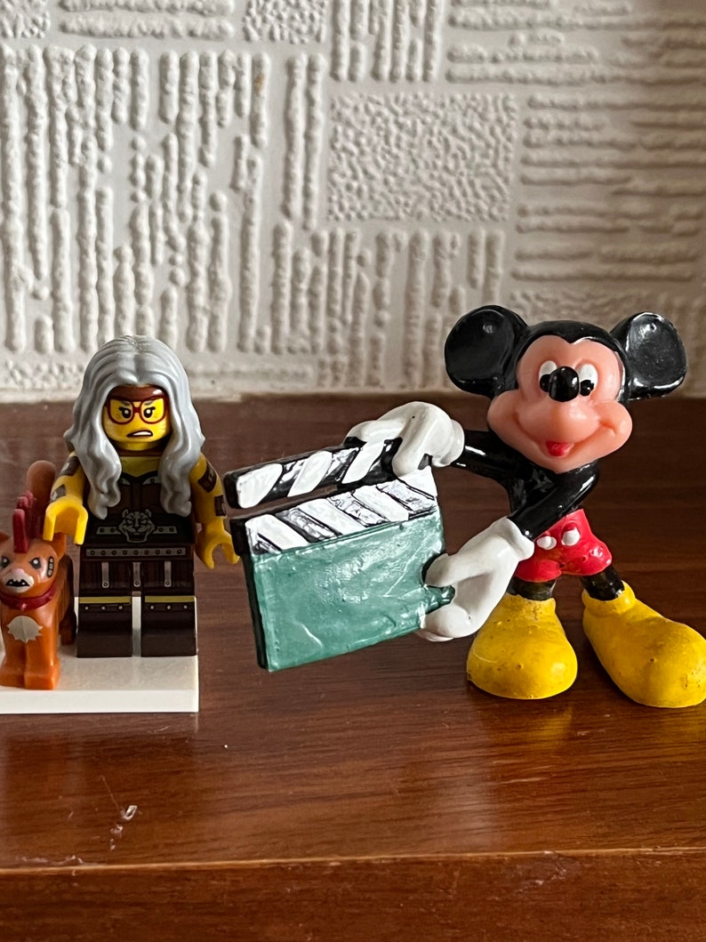 May include: A black and white clapperboard held by a red and yellow Mickey Mouse figurine. A Lego figurine with grey hair and a yellow and brown outfit stands next to Mickey Mouse.