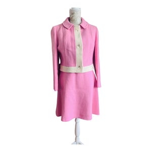 May include: A bright pink two-piece suit consisting of a long-sleeved jacket and a skirt. The jacket features a cream-colored vertical placket with three buttons and a matching cream-colored horizontal band at the waist. The fabric has a textured finish.