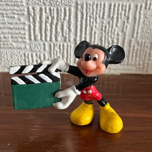 May include: A black and red plastic figurine of Mickey Mouse holding a green and white clapperboard.