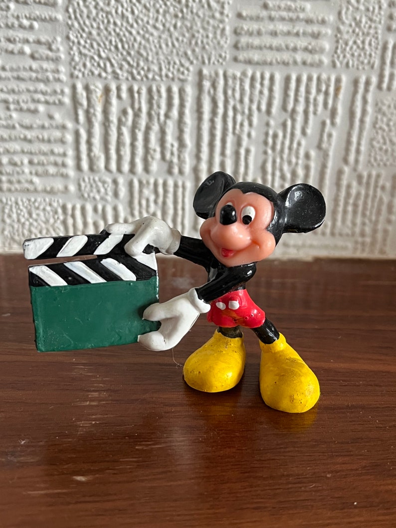 May include: A black and red plastic figurine of Mickey Mouse holding a green and white clapperboard.