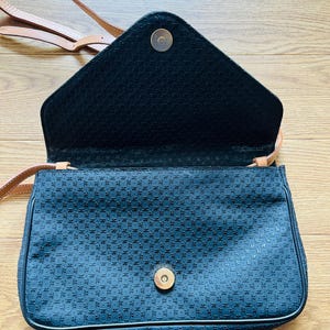 May include: A black crossbody bag with a geometric pattern and a tan strap. The bag has a flap closure with a gold-colored magnetic snap. The interior is black, and the bag appears to be made of a textured fabric.